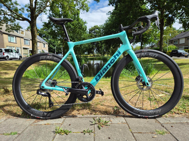 Een celeste Bianchi Infinito racefiets uitgerust met hoge, zwarte GreenGrav FLN-50 carbon wielen en een Shimano afmontage